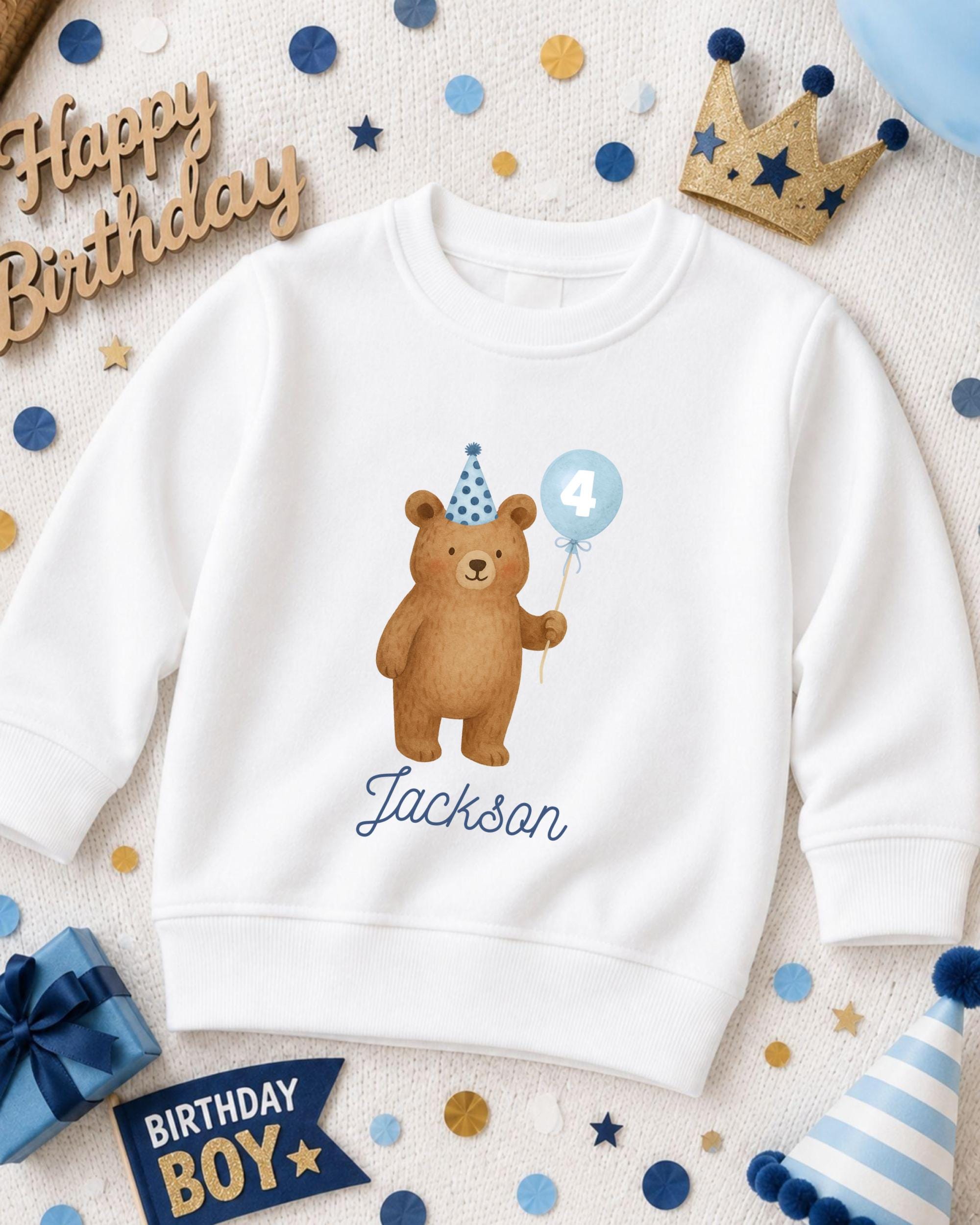 A white sweatshirt with a cartoon bear wearing a party hat and holding a blue balloon, surrounded by birthday decorations and a "Happy Birthday" sign.