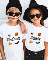 Two children wearing white t-shirts with Halloween-themed designs and the name "Hudson" and "Annie" printed on them, along with Halloween-themed icons such as bats, pumpkins, and skulls.