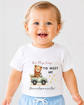 A smiling baby wearing a white t-shirt with a teddy bear and text that says "On My Way to Meet My Grandparents!"
