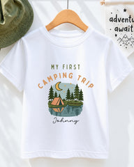 A white t-shirt with a graphic design depicting a camping scene with a tent, trees, and a lake, along with the text "My first camping trip" and "Johny" printed on it.