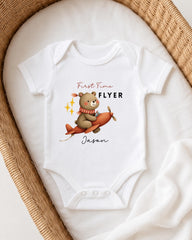 A white onesie with a cartoon image of a bear on an airplane, along with the text "First Time Flyer Jason" printed on it.