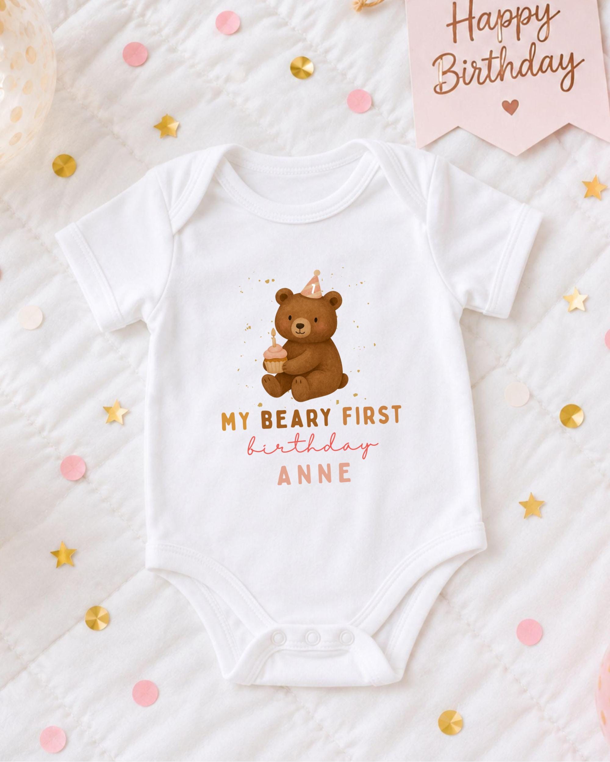 A white onesie with a teddy bear and text, placed on a bed with confetti and a birthday card.