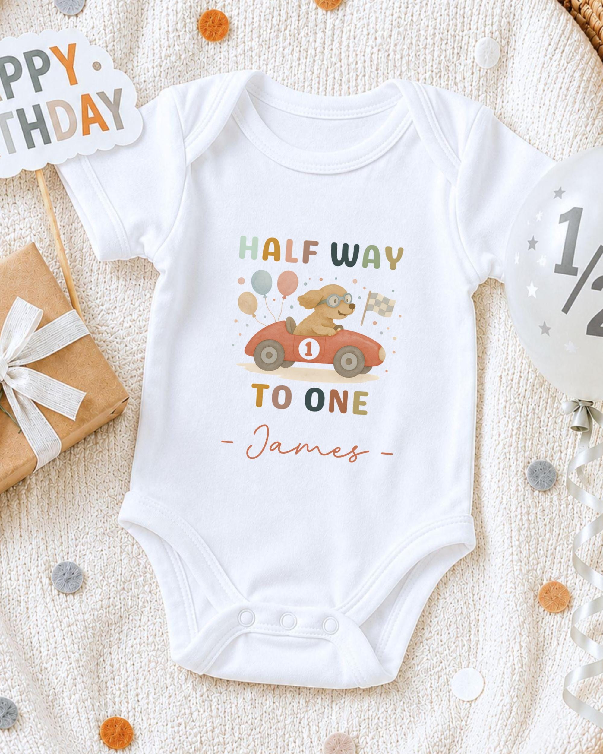 A white onesie with a cartoon image of a dog in a car and the text "Half Way to One - James" printed on it, along with a "Happy Birthday" sign and a gift box.