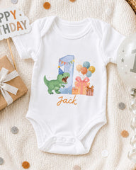 A white onesie with a dinosaur and birthday decorations, including a balloon and presents, is displayed on a white blanket.