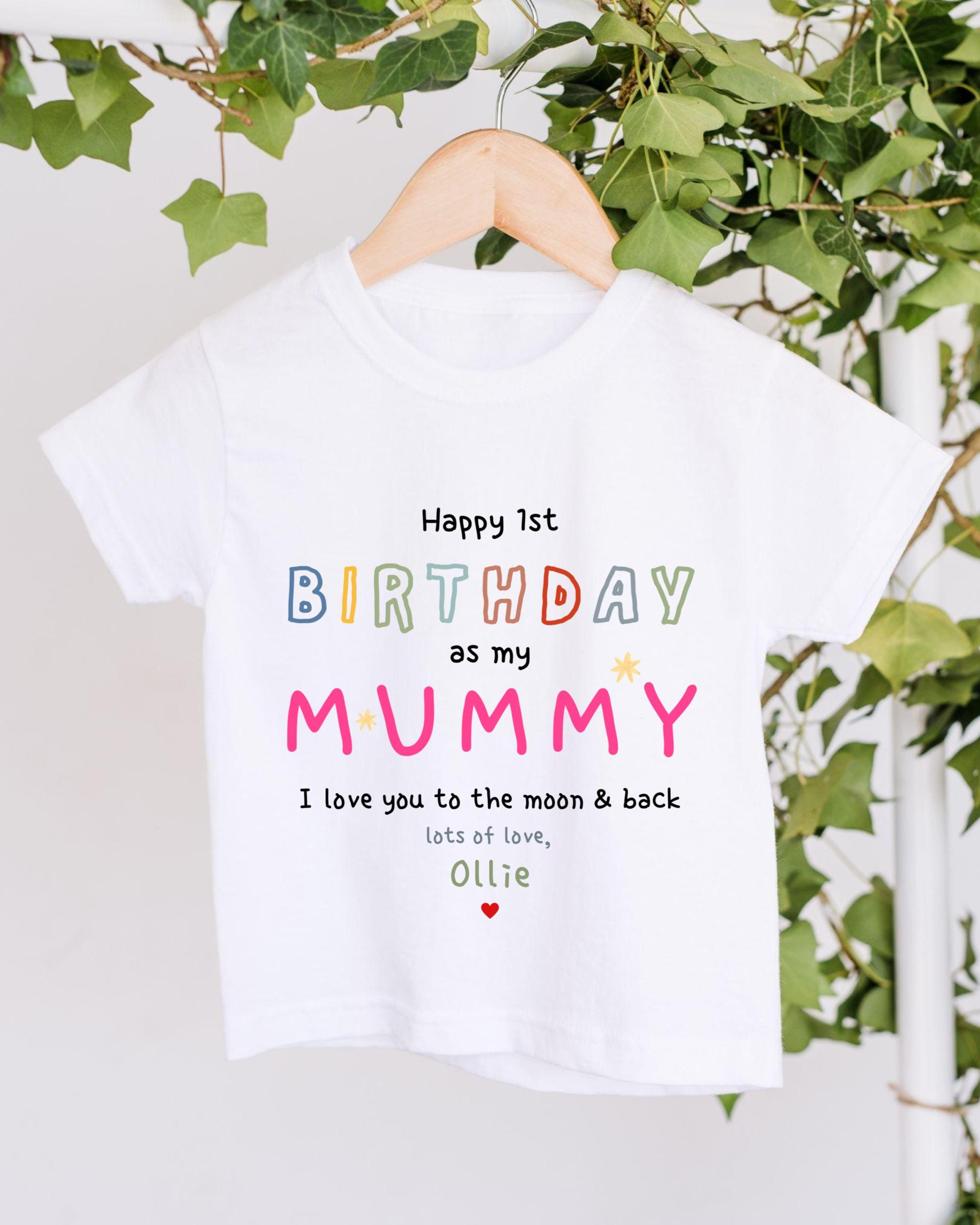A white t-shirt with a colorful birthday message for a mother hanging on a wooden hanger, with greenery in the background.
