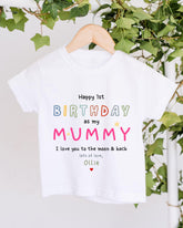 A white t-shirt with a colorful birthday message for a mother hanging on a wooden hanger, with greenery in the background.