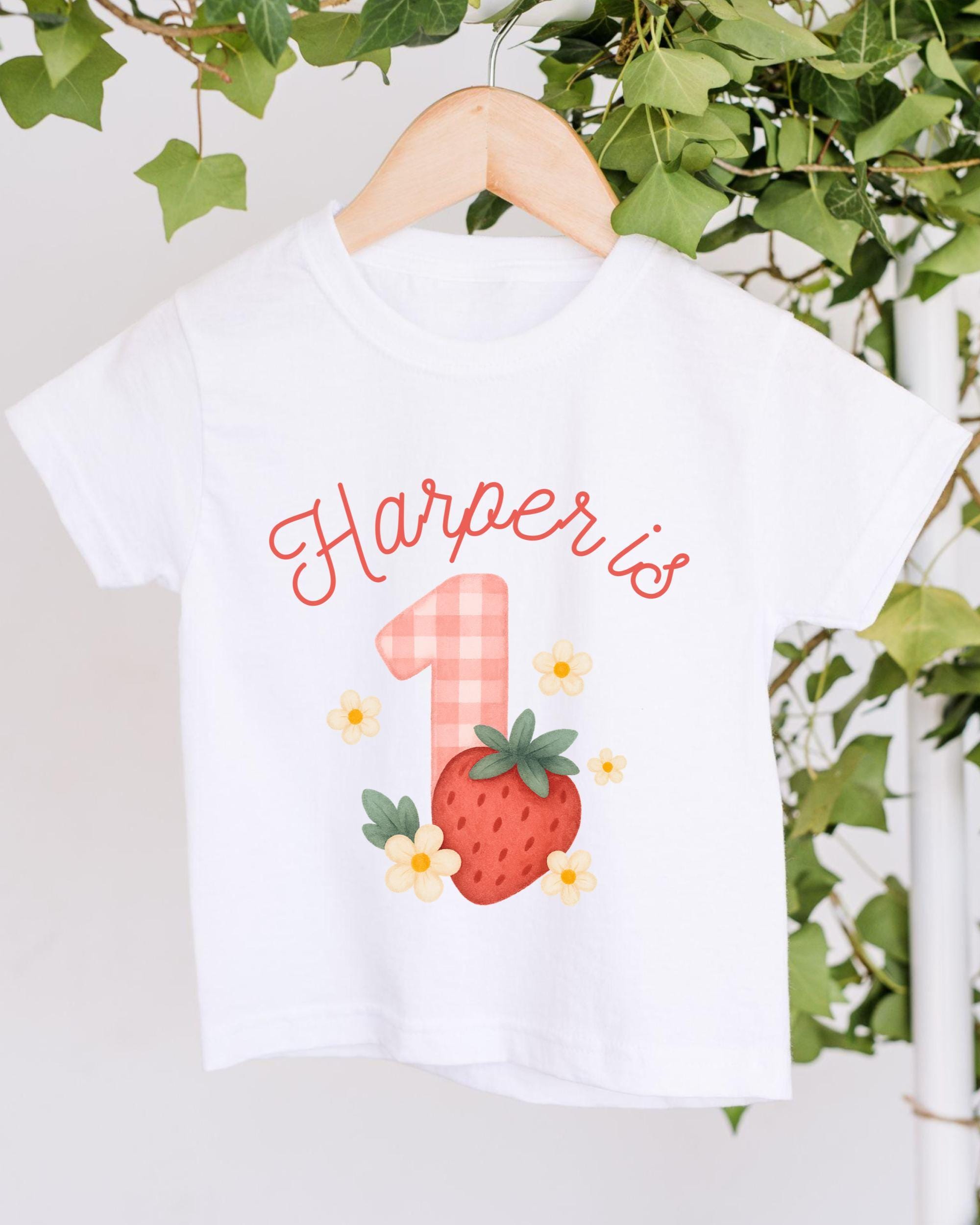 A white t-shirt with a personalized text and a strawberry graphic, hanging on a wooden hanger.