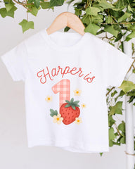 A white t-shirt with a personalized text and a strawberry graphic, hanging on a wooden hanger.