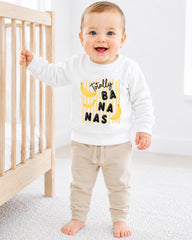 A smiling baby boy wearing a white sweatshirt with a banana graphic and the text "Totally BANANA" is standing next to a wooden crib.
