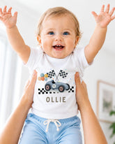 A smiling baby wearing a white t-shirt with a race car graphic and the name "Ollie" printed on it, being held up by someone's arms.