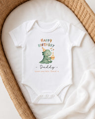 A white onesie with a cartoon dinosaur and the text "Happy Birthday Daddy" printed on it, placed in a woven basket.