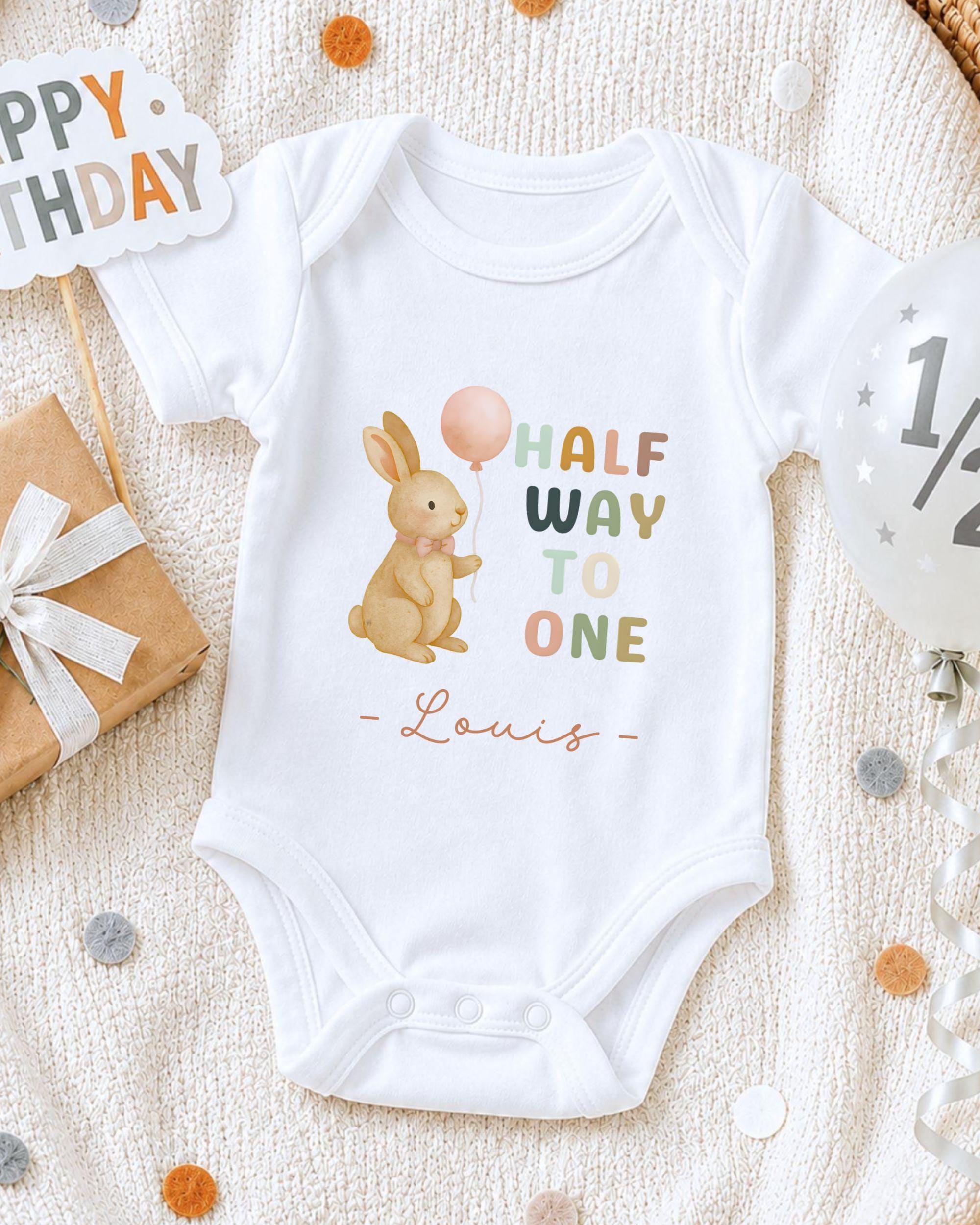 A white onesie with a cartoon rabbit and the text "Half Way to One" printed on it, along with the name "Louis" below the rabbit. The onesie is laid out on a white blanket, surrounded by birthday decorations such as a "Happy Birthday" sign, a balloon, and confetti.