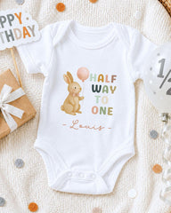 A white onesie with a cartoon rabbit and the text "Half Way to One" printed on it, along with the name "Louis" below the rabbit. The onesie is laid out on a white blanket, surrounded by birthday decorations such as a "Happy Birthday" sign, a balloon, and confetti.