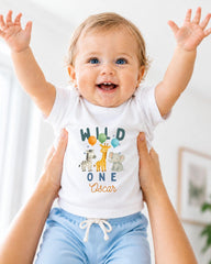 A smiling baby wearing a white t-shirt with a jungle animal design and the text "Wild One Oscar" is being held up by two people.