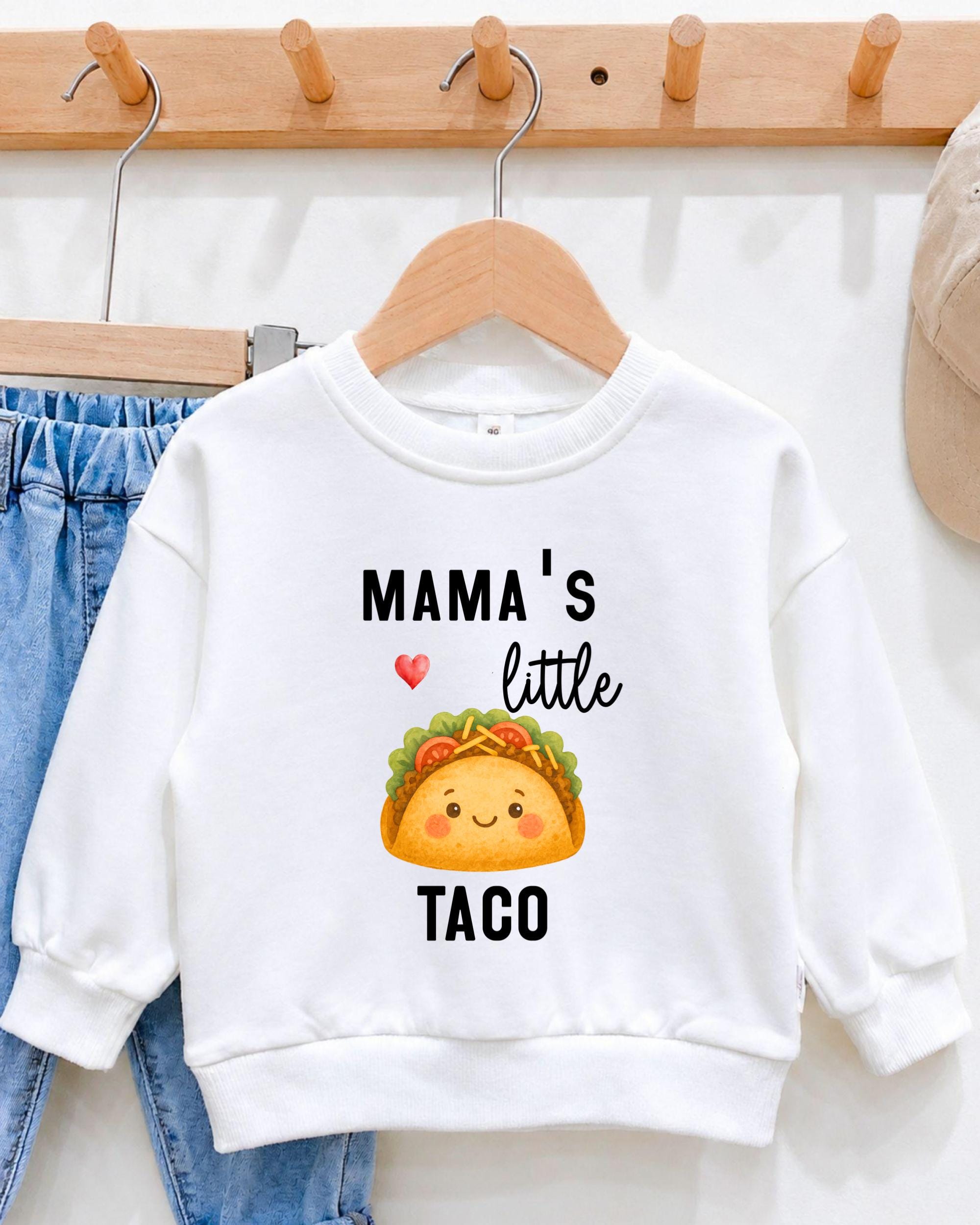 A white sweatshirt with a graphic of a taco and the text "Mama's little taco" printed on it, hanging on a wooden hanger.