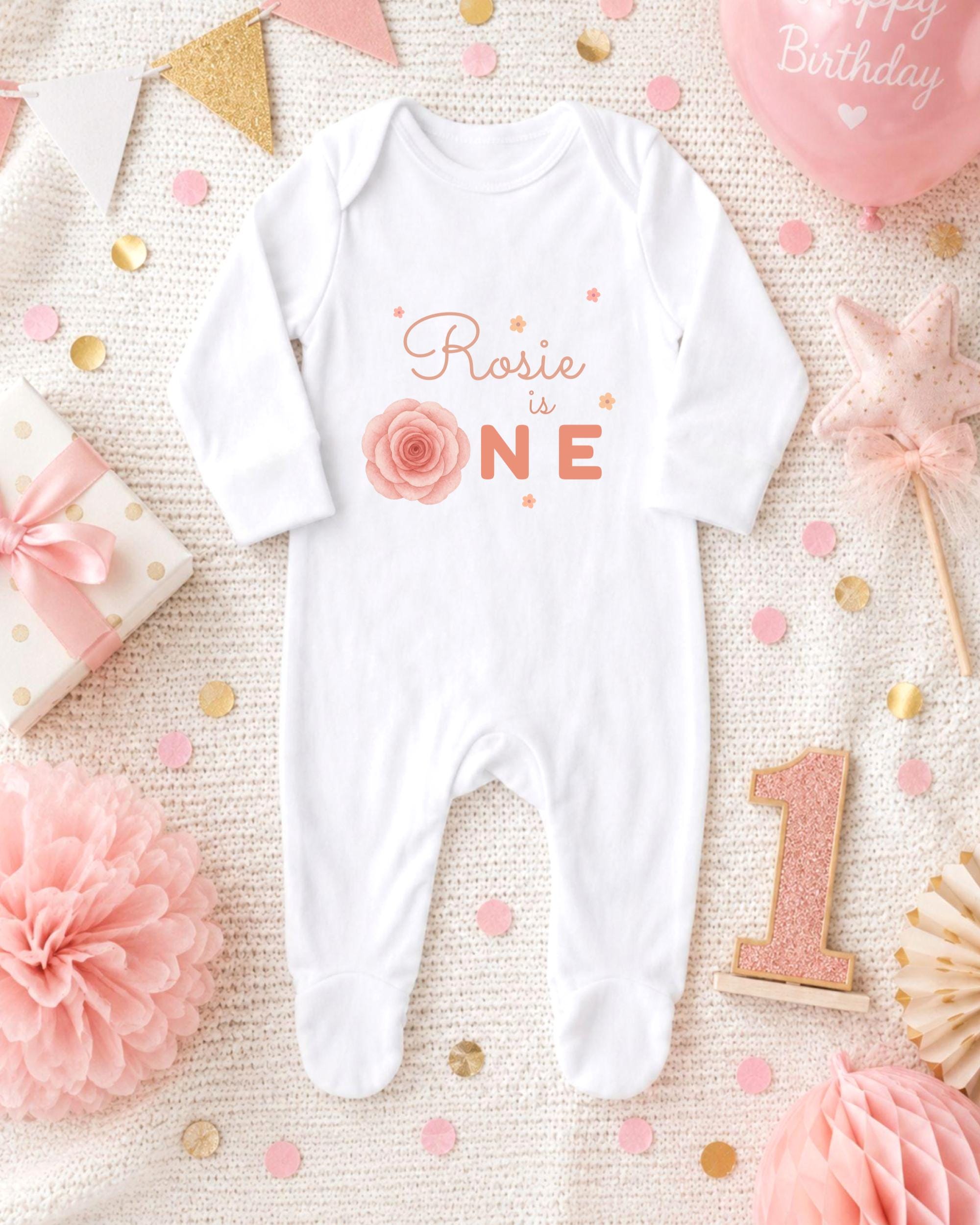 A white onesie with the text "Rosa is one" printed on it, surrounded by pink and gold decorations, confetti, and a pink balloon with the text "Happy Birthday" written on it.