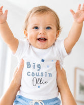 A smiling baby wearing a white shirt with the text "Big Cousin Ollie" is being held up by someone's arms.