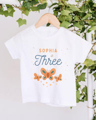 A white t-shirt with a design featuring butterflies and the text "Sophia is Three" printed on it, hanging on a wooden hanger.