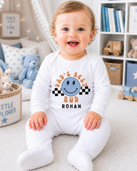 A smiling baby boy wearing a white onesie with a graphic and text, sitting on a carpeted floor in a room with a bookshelf and stuffed animals in the background.