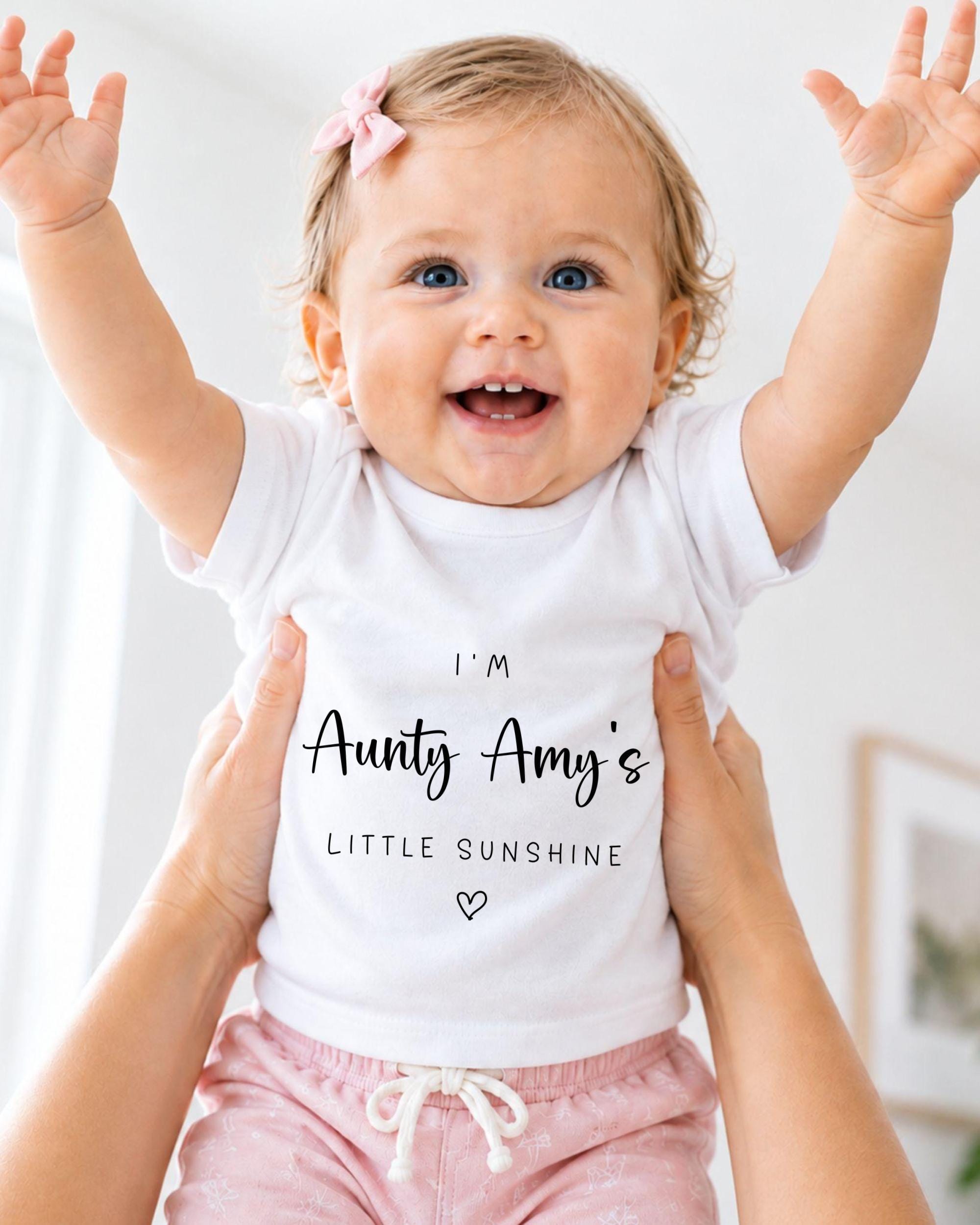 A smiling baby girl wearing a white t-shirt with text and a pink bow, being held up by someone's arms.
