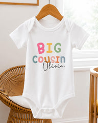 A white baby onesie with the text "BIG COUSIN Olivia" printed on it, hanging on a wooden hanger.