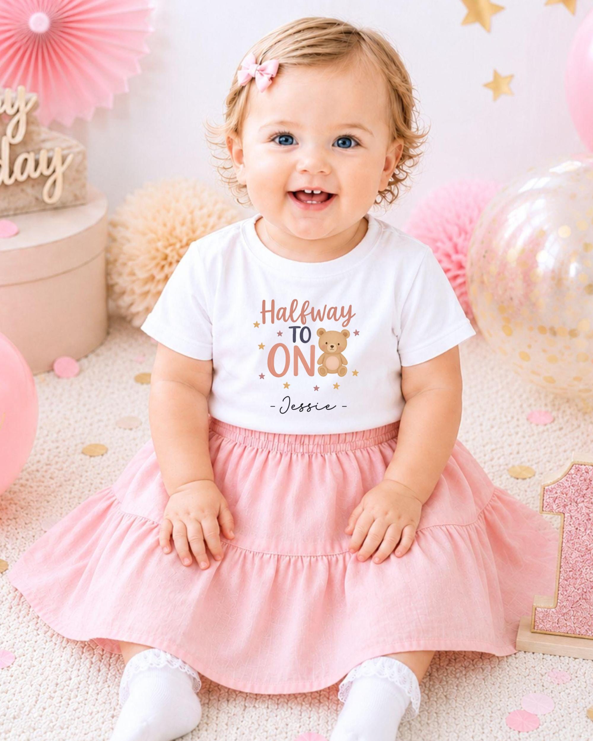 A smiling baby girl wearing a white t-shirt with a teddy bear and the text "Halfway to 1" sits on a pink and white background with pink and gold decorations.