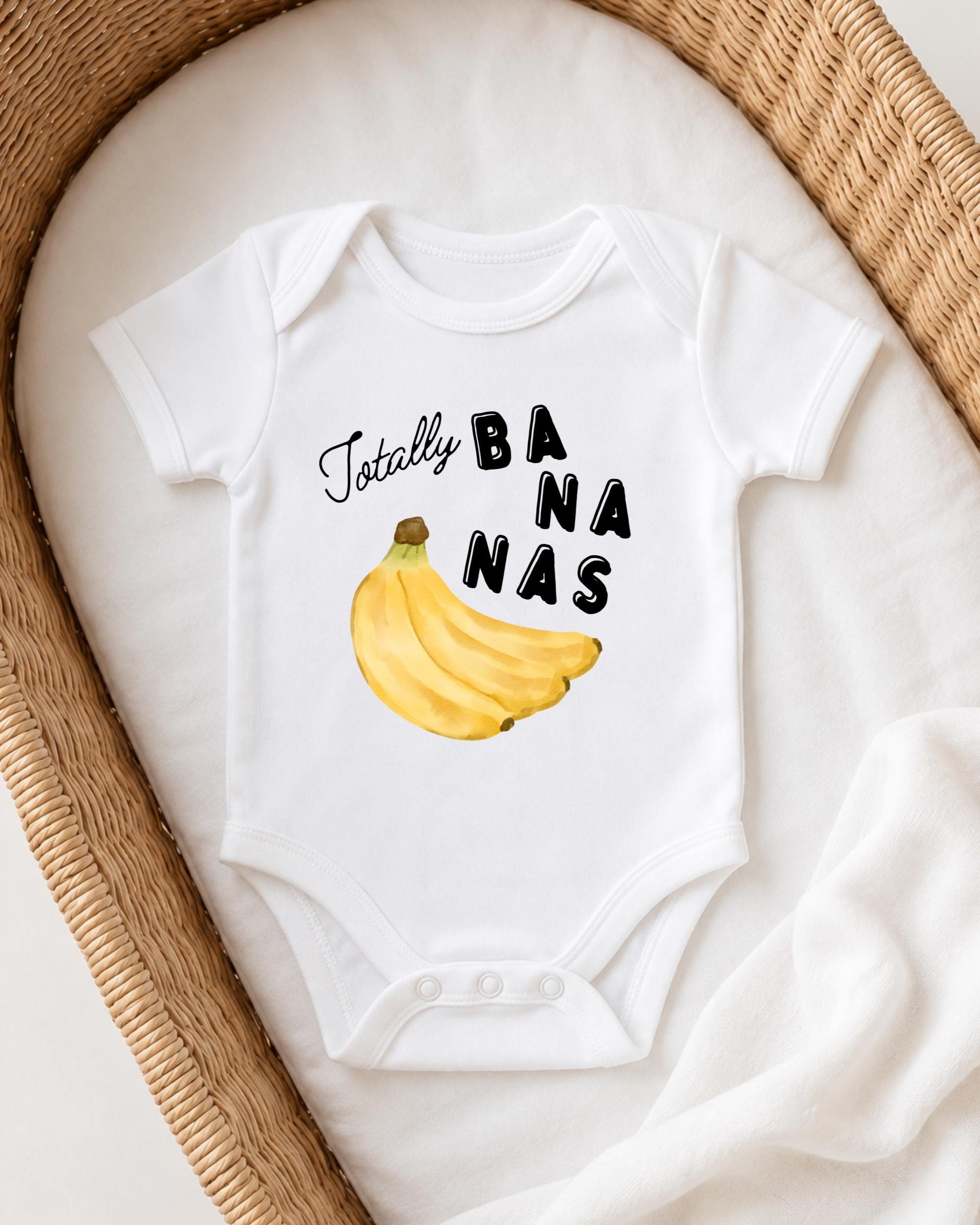A white onesie with a banana graphic and the text "Totally BANANA" printed on it, laid out on a white blanket.