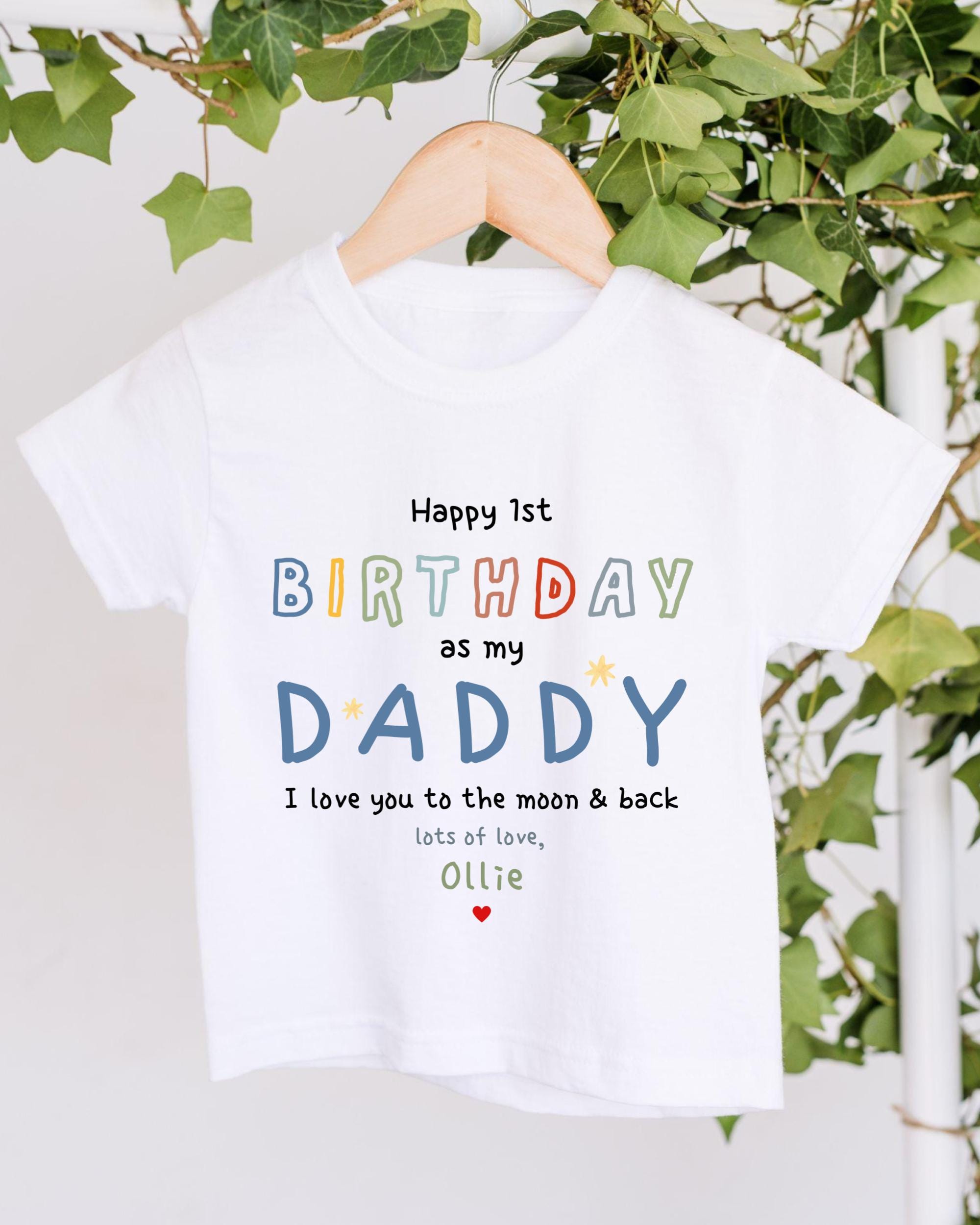 A white t-shirt with a birthday message printed on it, hanging on a wooden hanger.