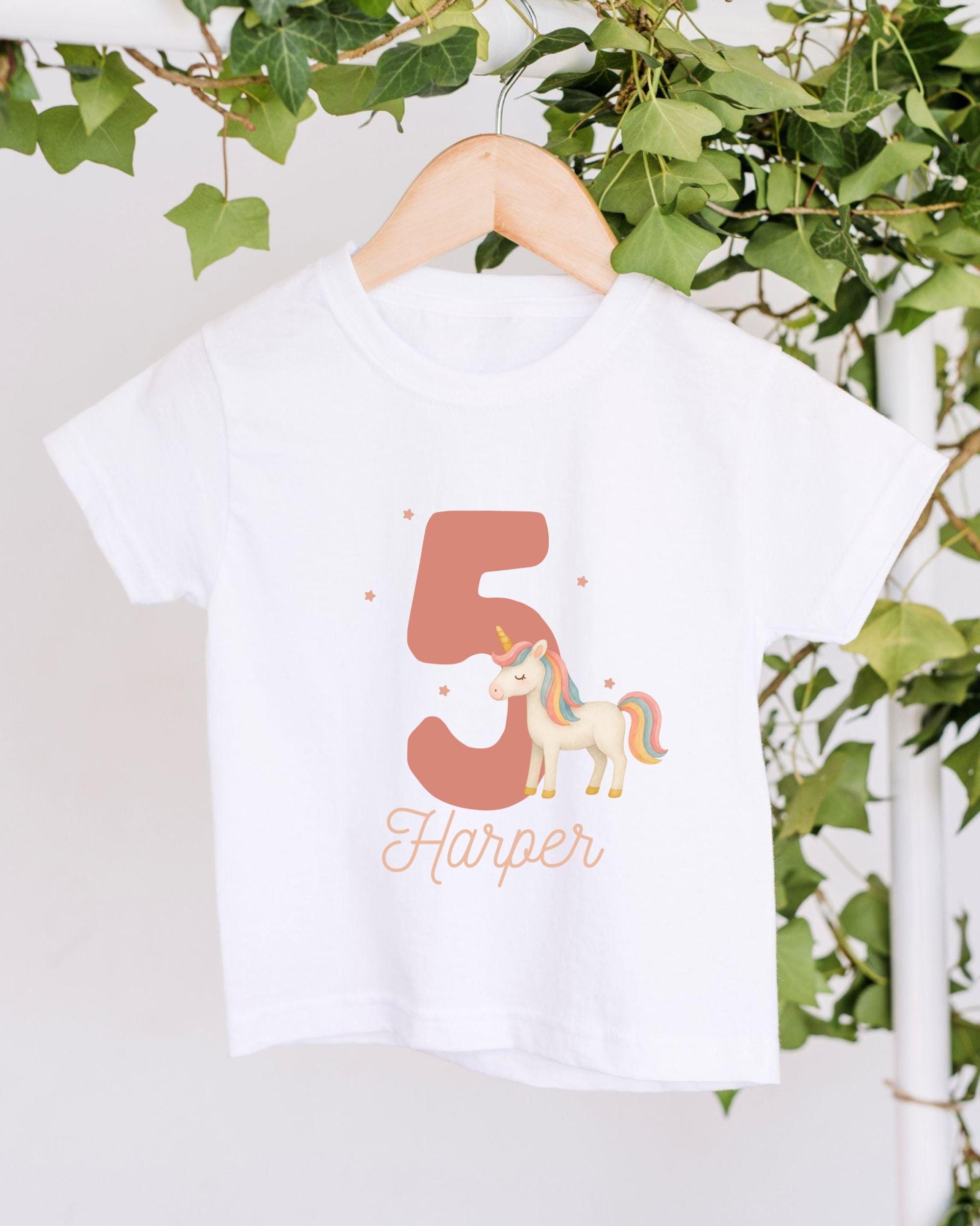 A white t-shirt with a unicorn graphic and the number 5 printed on it, hanging on a wooden hanger.