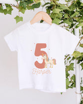 A white t-shirt with a unicorn graphic and the number 5 printed on it, hanging on a wooden hanger.