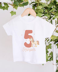 A white t-shirt with a unicorn graphic and the number 5 printed on it, hanging on a wooden hanger.