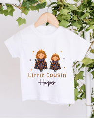 A white t-shirt with a graphic design featuring two cartoon princesses wearing crowns, surrounded by stars and vines. The text below the design reads "Little Cousin Harper".