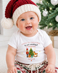 A smiling baby wearing a red and white Santa hat and a white t-shirt with a Christmas-themed design, sitting in front of a Christmas tree.