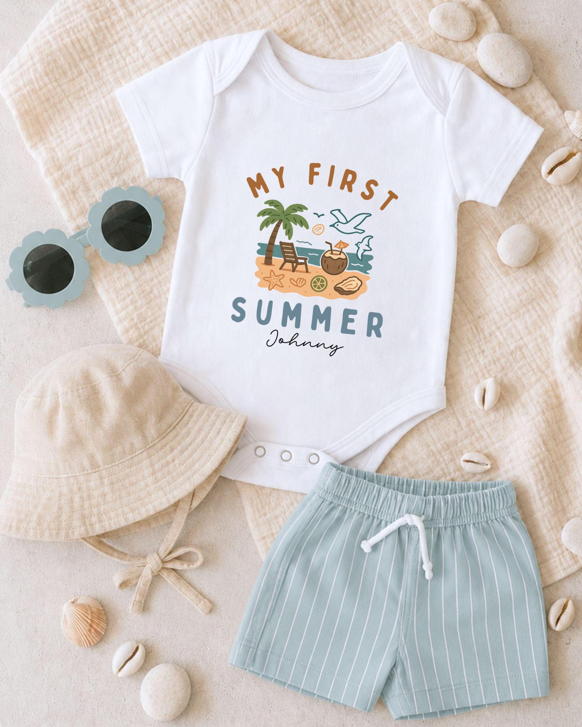 A baby's outfit, including a white onesie with a summer theme, a beige bucket hat, and light blue shorts, along with sunglasses and seashells, are arranged on a beige surface.