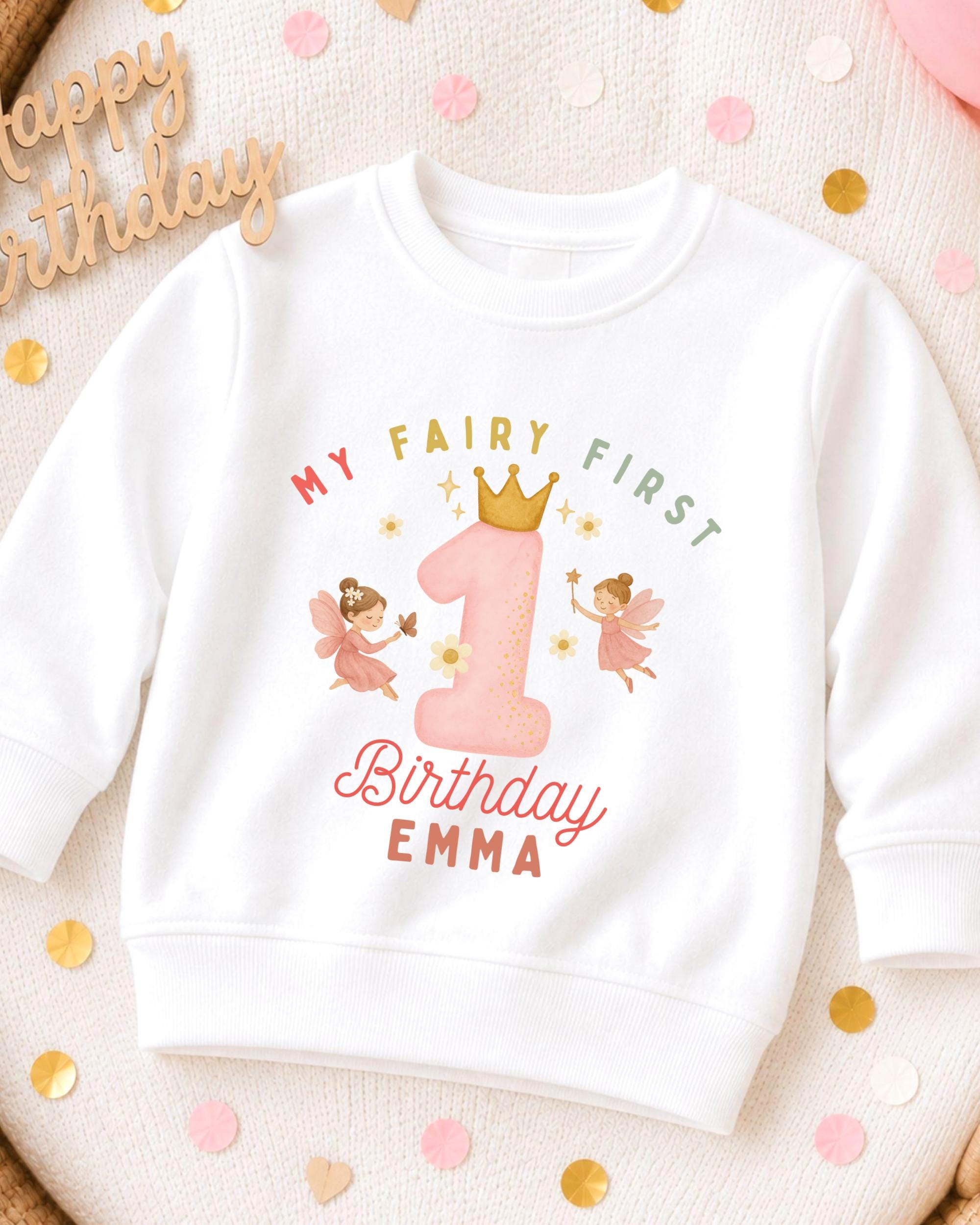 A white sweatshirt with a personalized birthday design for a child named Emma. The design features a large number one with a crown, surrounded by two pink fairy characters. The text on the sweatshirt reads "My Fairy First Birthday Emma".
