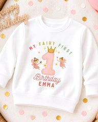 A white sweatshirt with a personalized birthday design for a child named Emma. The design features a large number one with a crown, surrounded by two pink fairy characters. The text on the sweatshirt reads "My Fairy First Birthday Emma".