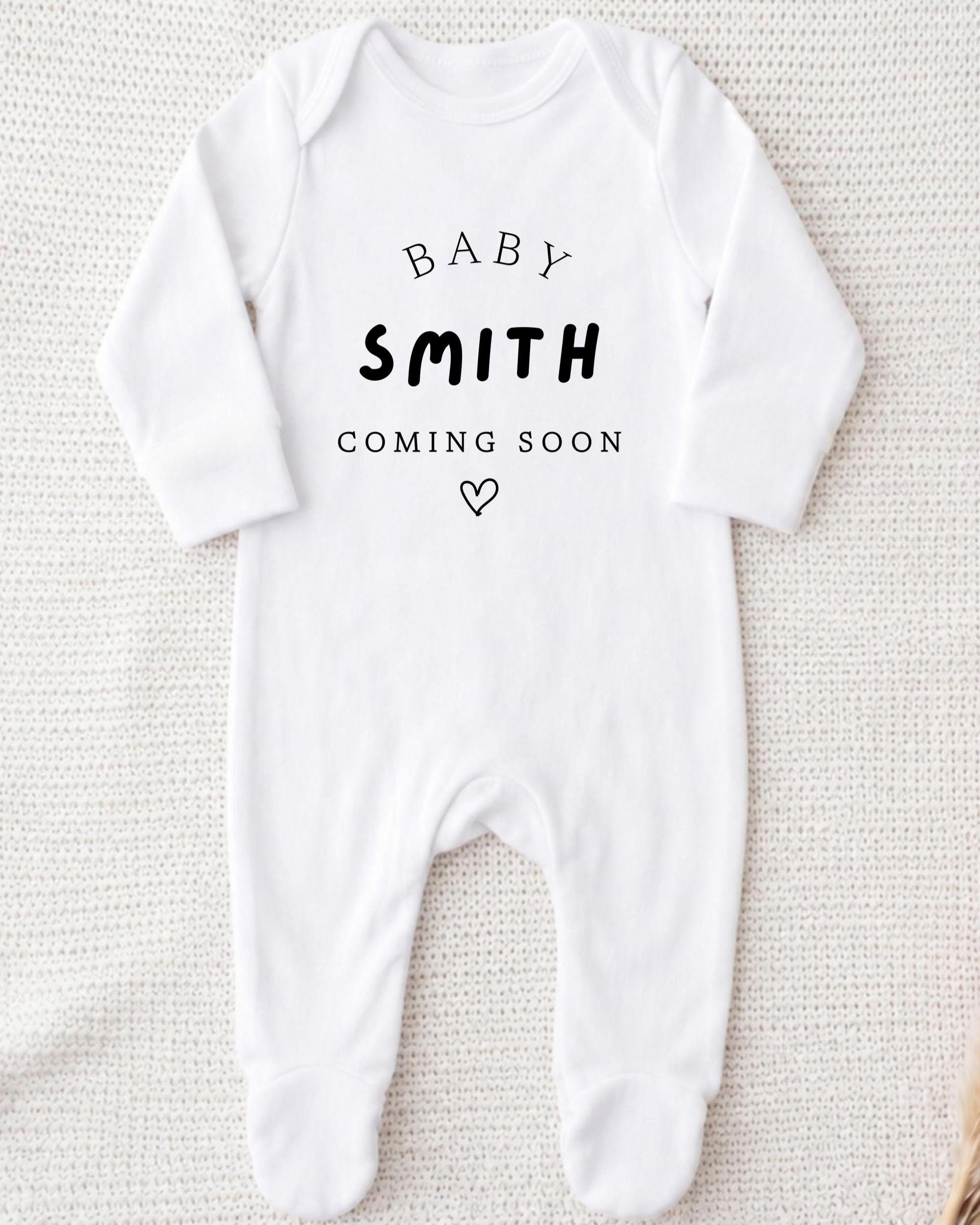 A white onesie with the text "BABY SMITH COMING SOON" printed on it.