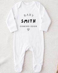 A white onesie with the text "BABY SMITH COMING SOON" printed on it.