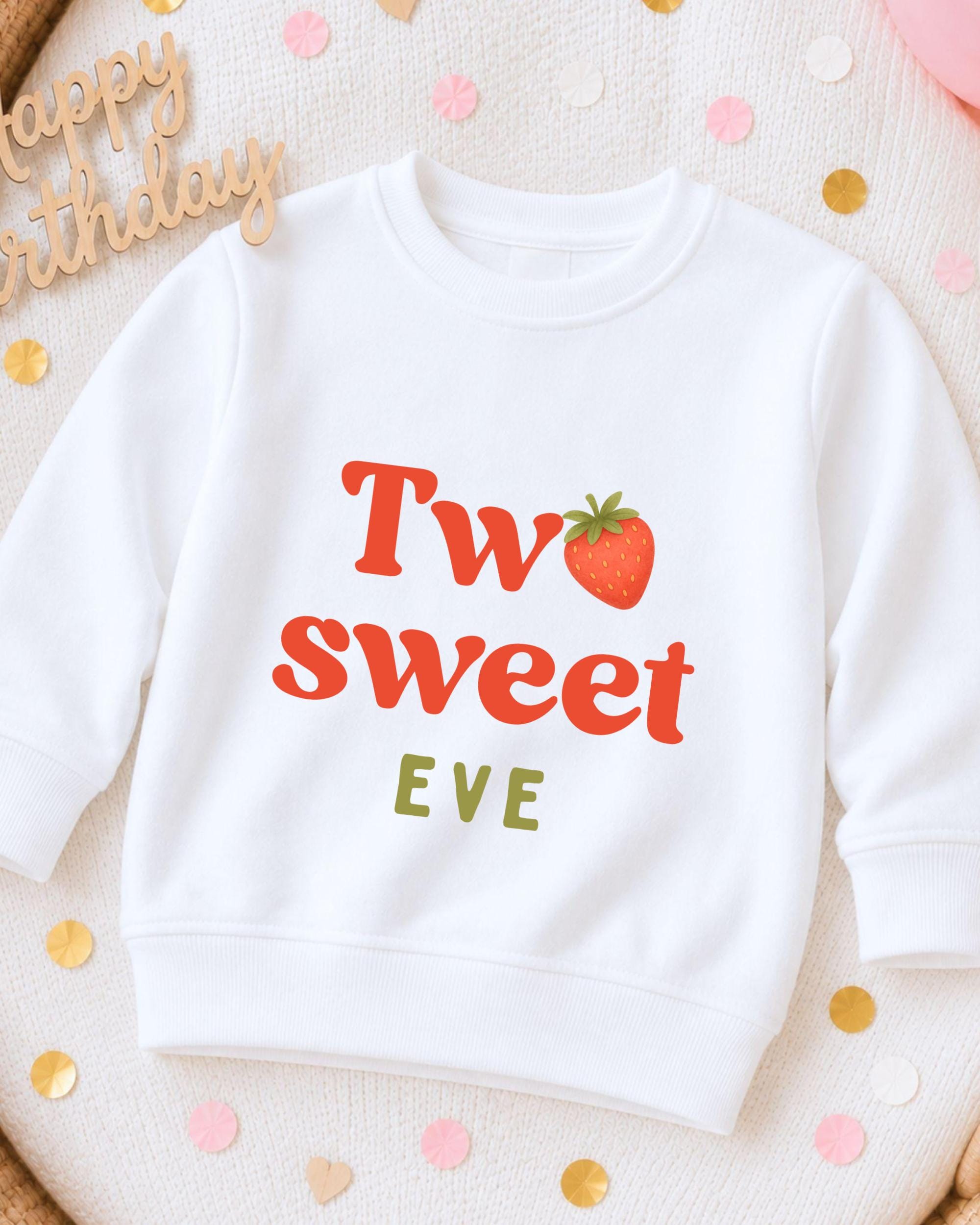 A white sweatshirt with a red strawberry graphic and the text "Two sweet EVE" printed on it, surrounded by confetti.