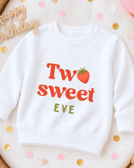 A white sweatshirt with a red strawberry graphic and the text "Two sweet EVE" printed on it, surrounded by confetti.