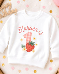 A white sweatshirt with a personalized design on it, featuring a strawberry and flowers, and the name "Harper's" written on it.