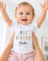 A smiling baby girl wearing a white t-shirt with the text "BIG SISTER Amelia EST 2023" is being held up by someone's arms.