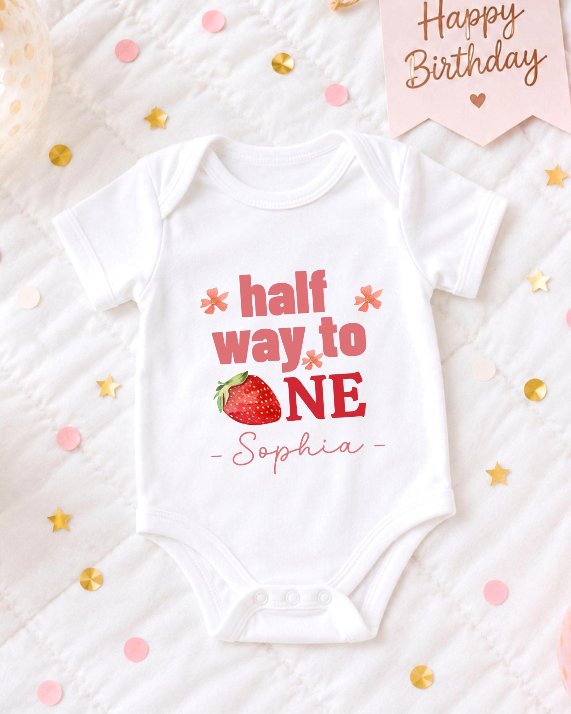 A white onesie with the text "half way to one" printed on it, along with a strawberry image, is displayed on a bed surrounded by confetti.