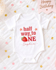A white onesie with the text "half way to one" printed on it, along with a strawberry image, is displayed on a bed surrounded by confetti.