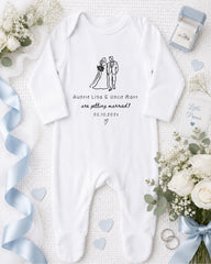A white onesie with a graphic of a bride and groom, along with text that reads "Auntie Lisa & Uncle Matt are getting married! 20.10.2023", is displayed on a white background. Surrounding the onesie are various wedding-related items, including a ring box, a bouquet of white flowers, and a blue ribbon.