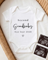 A white onesie with the text "Second Grandbaby Due Sept 2028" printed on it, along with a heart symbol, placed on a bed next to two black and white images of an Ultrasound scan.