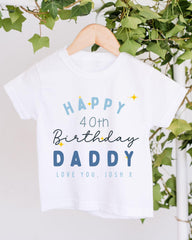 A white t-shirt with a birthday message printed on it, hanging on a wooden hanger.