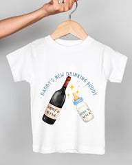 A white t-shirt with a graphic design depicting a wine bottle and a baby bottle, along with the text "DADDY'S NEW DRINKING BUDDY" and "JOSH'S MILK".