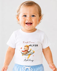 A smiling baby wearing a white t-shirt with a cartoon image of a teddy bear and the text "First Time Flyer Ashley" printed on it.