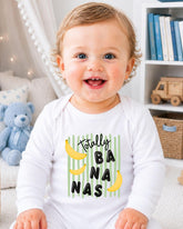 A smiling baby wearing a white onesie with a graphic of bananas and the text "Totally BANANA" is sitting in a room with a teddy bear and bookshelf in the background.